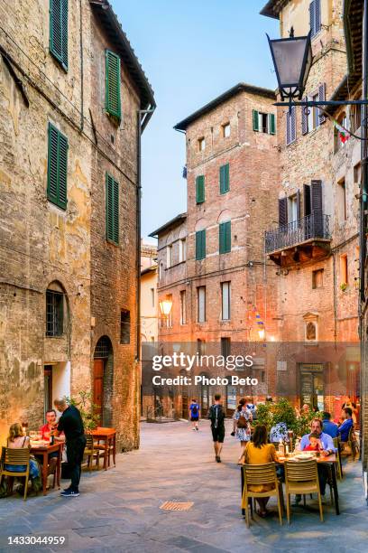 some tourists enjoy life in a restaurant along a beautiful stone alley in the medieval heart of siena in tuscany - siena italy stockfoto's en -beelden