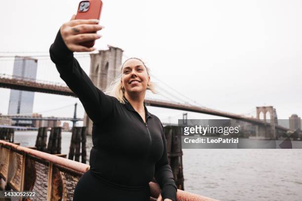 happy woman takes a selfie in front of brooklyn bridge in new york during her running session - new sport content stock pictures, royalty-free photos & images