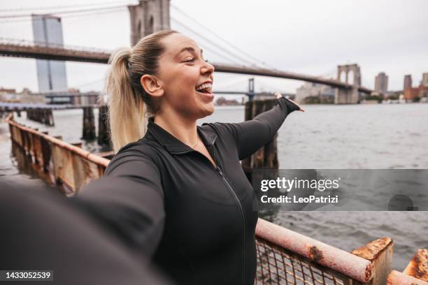 happy woman takes a selfie in front of brooklyn bridge in new york during her running session - new sport content stock pictures, royalty-free photos & images