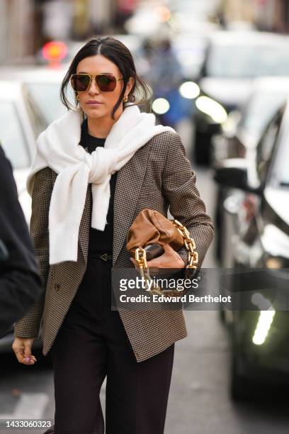 Guest wears brown sunglasses, gold earrings, a black t-shirt, a white wool knot at the shoulder pullover, a brown and beige houndstooth print pattern...