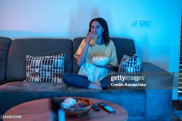 young latin female drinks orange juice while watching tv. she holds popcorn - netflix stock pictures, royalty-free photos & images