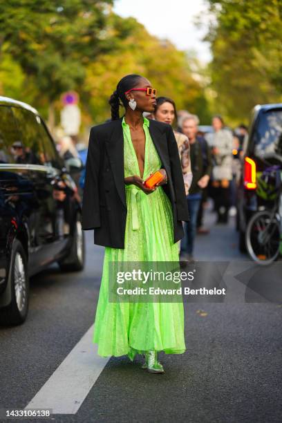 Guest wears red and orange sunglasses, rhinestones large heart pendant earrings, a neon green lace print pattern V-neck / pleated long dress, a black...