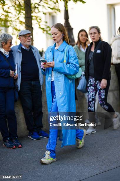 Guest wears gold earrings, a white pearls necklace, necklaces, a blue shiny varnished leather long coat, a pale blue shiny leather shoulder bag, a...