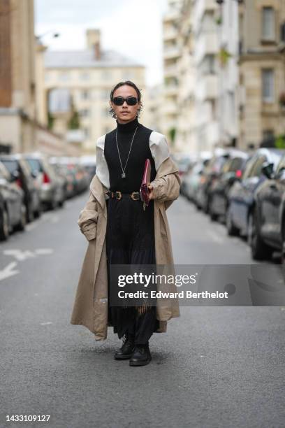 Guest wears black sunglasses, a silver chain necklace, a black turtleneck pullover, a black shiny leather with gold buckle belt, a beige long coat, a...