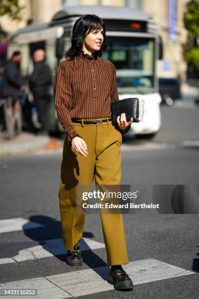 Guest wears a brown striped print pattern high neck ruffled blouse, a black shiny leather small belt, a black shiny leather braided clutch, rings,...