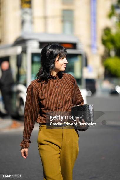 Guest wears a brown striped print pattern high neck ruffled blouse, a black shiny leather small belt, a black shiny leather braided clutch, rings,...