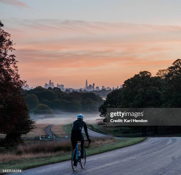 an unrecognisable cyclist on a winding road at dawn in a london park - city of london in the background - richmond upon thames stockfoto's en -beelden