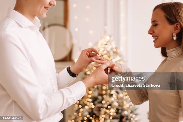 a guy in love makes a marriage proposal to his girlfriend on christmas day in the luxury richly decorated interior of the living room against the background of christmas trees at home. the groom puts an engagement ring on the bride's hand - winter bride stock pictures, royalty-free photos & images