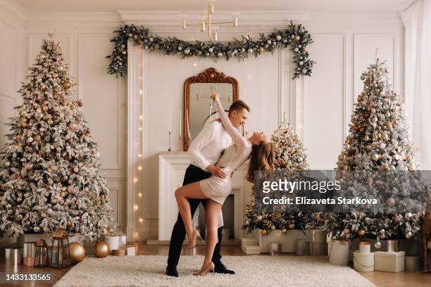 a couple in love in elegant fashionable outfits embrace, rejoice and celebrate the christmas holiday in a bright luxury  decorated interior with christmas trees in the living room of the house. a young man in a suit and a woman in a dress - fashion designer married hugging bildbanksfoton och bilder