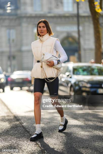 Guest wears logo earrings, a white latte high neck / sleeveless jacket from Ganni, a black shiny leather belt from Chanel, a white wool pullover, a...