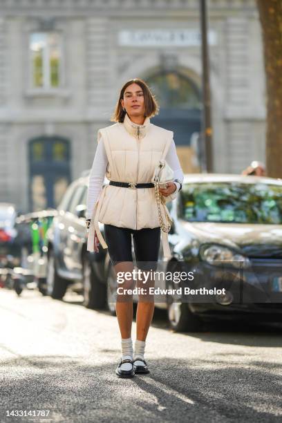 Guest wears logo earrings, a white latte high neck / sleeveless jacket from Ganni, a black shiny leather belt from Chanel, a white wool pullover, a...