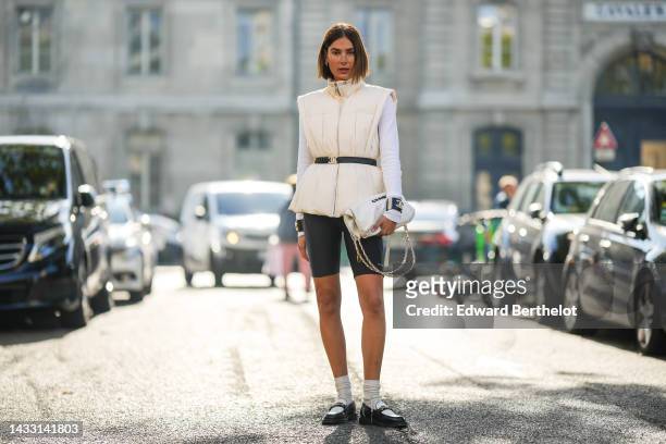 Guest wears logo earrings, a white latte high neck / sleeveless jacket from Ganni, a black shiny leather belt from Chanel, a white wool pullover, a...