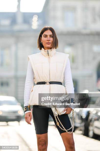 Guest wears logo earrings, a white latte high neck / sleeveless jacket from Ganni, a black shiny leather belt from Chanel, a white wool pullover, a...