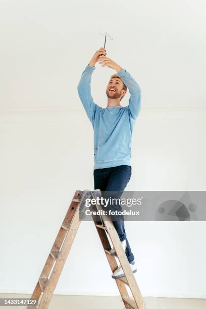 joven de pie en la escalera y cambiando la bombilla en casa - escalera plegable fotografías e imágenes de stock