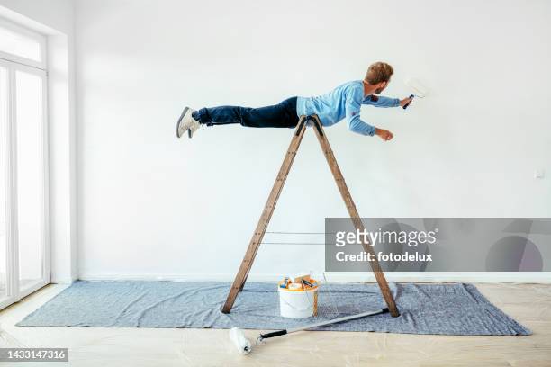 young man balancing on a ladder and painting living room wall - humor stockfoto's en -beelden