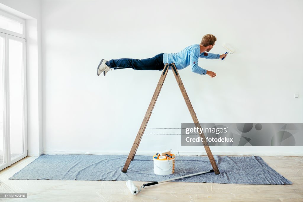Young man balancing on a ladder and painting living room wall