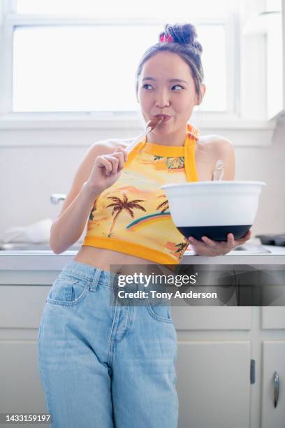 young woman in kitchen holding mixing bowl and licking batter off of spoon - crop top stock pictures, royalty-free photos & images