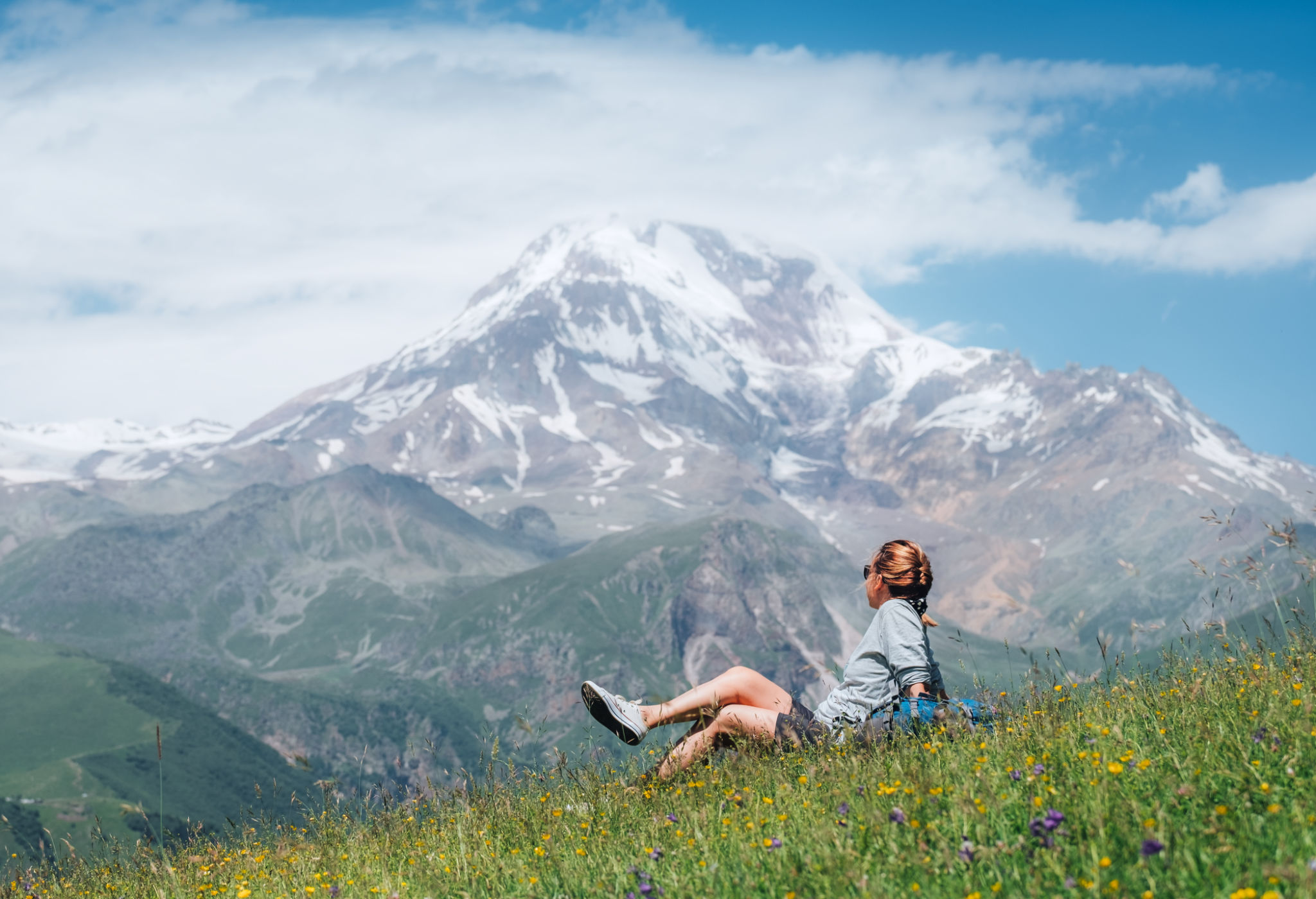 Backpacker woman sitting on a green grass hill and enjoying snowy slopes of Kazbek 5054m mountain with a backpack while she walking by green grass hill. East Caucasus mountains, Georgia. Backpacker woman sitting on a green grass hill and enjoying snowy slopes of Kazbek 5054m mountain with a backpack while she walking by green grass hill. East Caucasus mountains, Georgia.