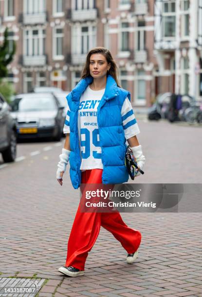 Talissa Benamou wears red pants, football shirt jersey with print, blue puffer vest, creme white gloves, clutch with print, Converse shoes on October...
