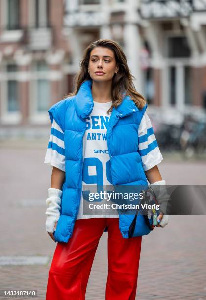 Talissa Benamou wears red pants, football shirt jersey with print, blue puffer vest, creme white gloves, clutch with print, Converse shoes on October...