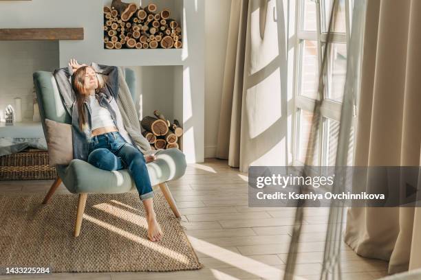 young woman at home sitting on modern chair in front of window relaxing in her living room reading book and drinking coffee or tea - man woman comfort stock-fotos und bilder