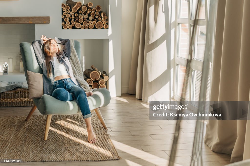 Young woman at home sitting on modern chair in front of window relaxing in her living room reading book and drinking coffee or tea
