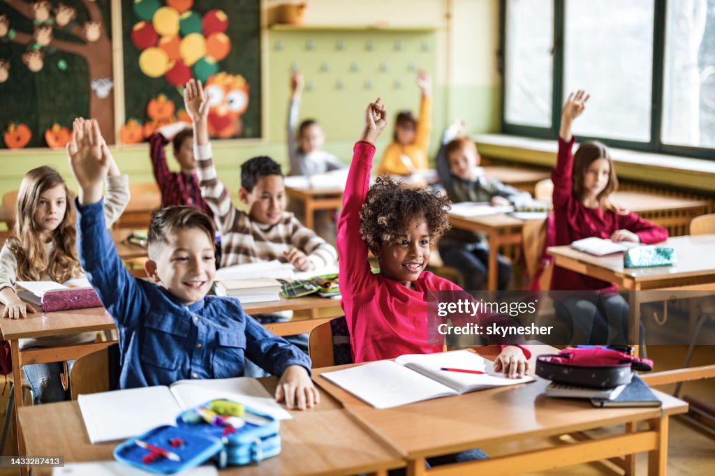 Happy elementary students raising their hands on a class at school.