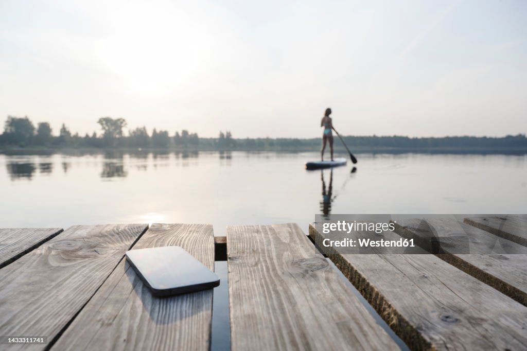 Mobile phone on jetty with woman doing standup paddleboarding in background at sunset