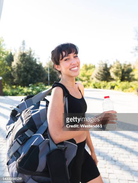 happy woman with gym bag and water bottle walking on footpath - gym bag stock-fotos und bilder