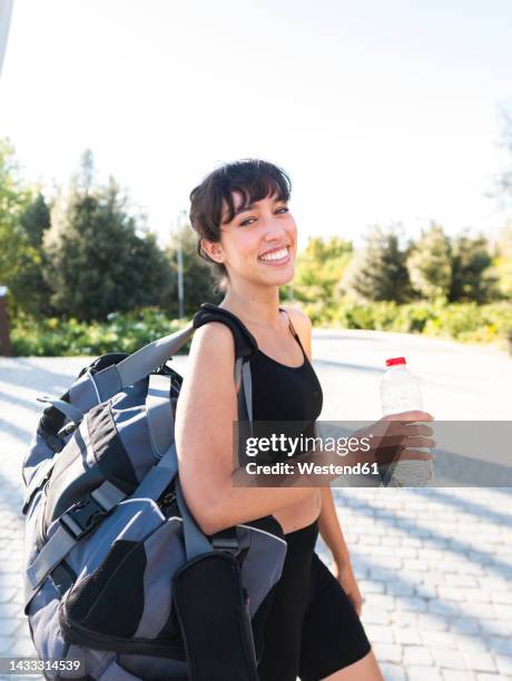 happy woman with gym bag and water bottle walking on footpath - carrying sports bag foto e immagini stock