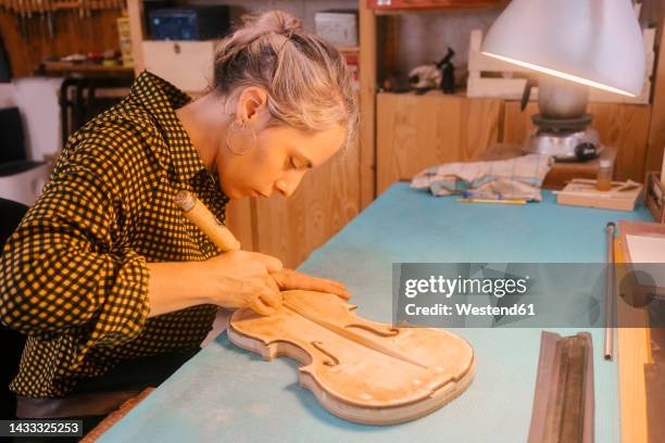 luthier with hand tool carving on violin at desk in workshop - instrument maker fotografías e imágenes de stock