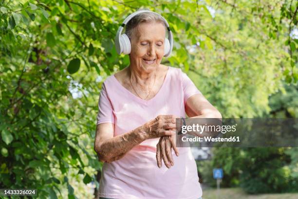 smiling senior woman with smart watch listening to music through headphones in park - smart watch stock pictures, royalty-free photos & images