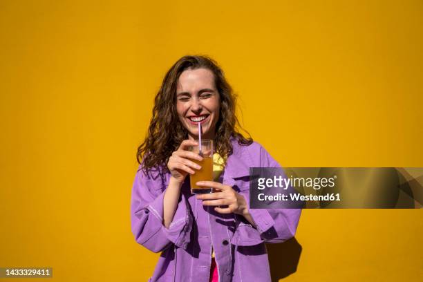 cheerful woman with eyes closed drinking juice in front of yellow wall - chaqueta púrpura fotografías e imágenes de stock