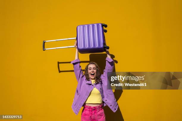 excited woman lifting wheeled luggage in front of yellow wall - soulever photos et images de collection