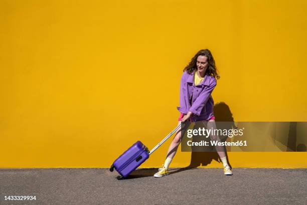 woman pulling wheeled luggage in front of yellow wall - rullväska bildbanksfoton och bilder
