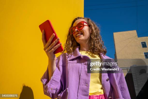 happy woman text messaging through smart phone near wall on sunny day - viewpoint stock pictures, royalty-free photos & images