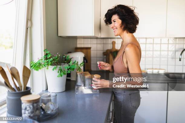 smiling woman holding jar looking through window standing at kitchen counter - jars kitchen stock pictures, royalty-free photos & images