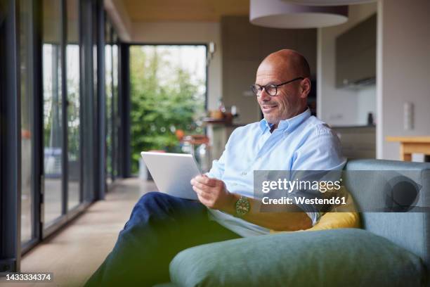 smiling man using tablet pc sitting on sofa at home - westend61 fotografías e imágenes de stock