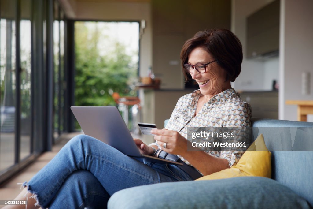Happy woman paying with credit card through laptop on sofa at home