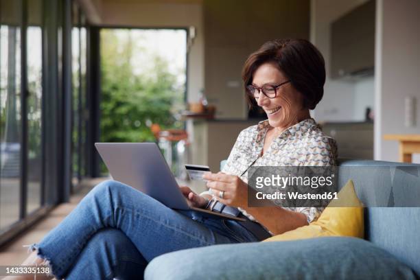 happy woman paying with credit card through laptop on sofa at home - silver surfer stock pictures, royalty-free photos & images