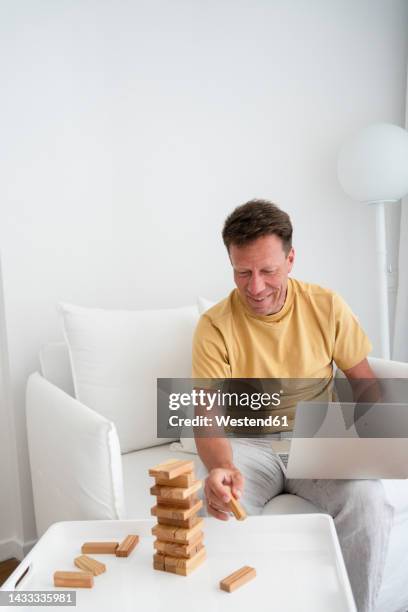 happy mature with laptop and toy blocks sitting in armchair at home - jenga stockfoto's en -beelden