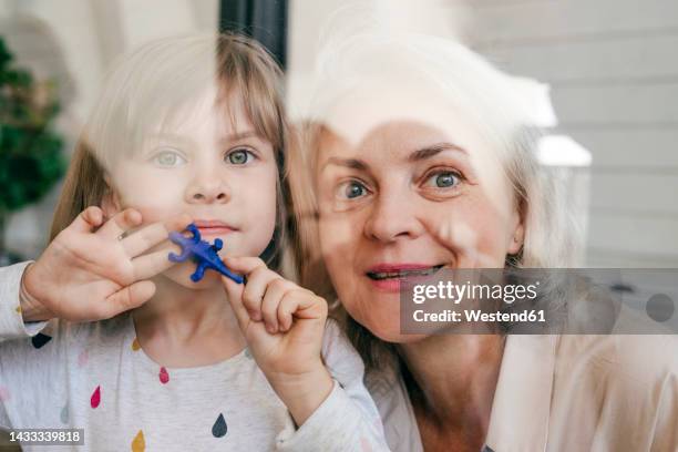 thoughtful grandmother and granddaughter with toy looking out of window - granddaughter stock pictures, royalty-free photos & images