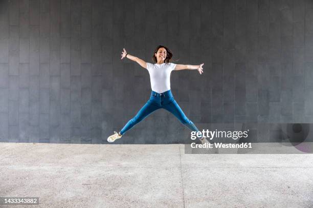 happy woman with arms outstretched jumping on footpath in front of gray wall - legs apart stock pictures, royalty-free photos & images