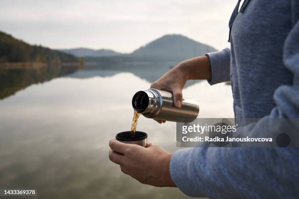 pouring warm tea from a vacuum bottle into a cup during a tourist trip at a lake - flask imagens e fotografias de stock