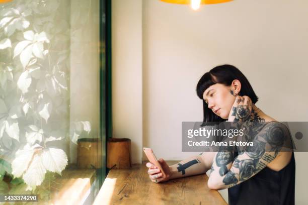 beautiful extraordinary woman with many tattoos and short dark hair is using smartphone sitting at a wooden table against window with many greens outside. she is wearing a black dress. concept of appearance that goes out of the frames - dress short sleeve stockfoto's en -beelden