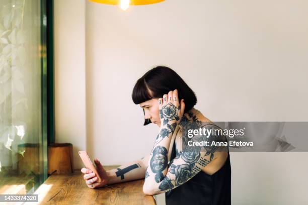 beautiful extraordinary woman with many tattoos and short dark hair is using smartphone sitting at a wooden table against window. she is wearing a black dress. concept of appearance that goes out of the frames - dress short sleeve stockfoto's en -beelden