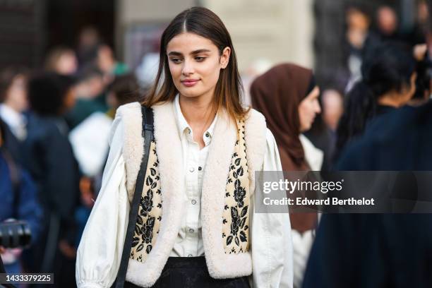 Taylor Hill wears gold earrings, a white denim shirt, a beige with black embroidered flower print pattern with sheep interior sleeveless gilet, black...