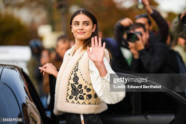 Taylor Hill wears gold earrings, a white denim shirt, a beige with black embroidered flower print pattern with sheep interior sleeveless gilet, black...