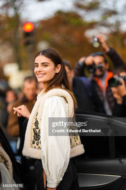 Taylor Hill wears gold earrings, a white denim shirt, a beige with black embroidered flower print pattern with sheep interior sleeveless gilet, black...