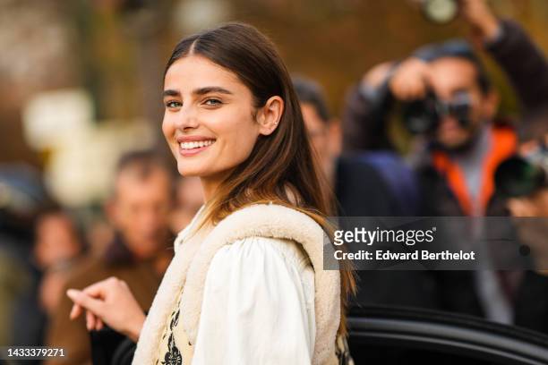 Taylor Hill wears gold earrings, a white denim shirt, a beige with black embroidered flower print pattern with sheep interior sleeveless gilet, black...